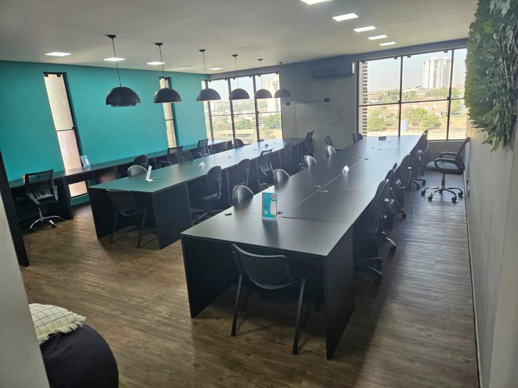 Estações de trabalho compartilhadas do coworking Trinova: mesas pretas em fileira, cadeiras e janelas com vista para a cidade